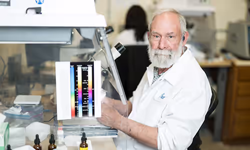 Senior man in a white lab coat at a laboratory bench, operating a scientific instrument with a color reference strip.