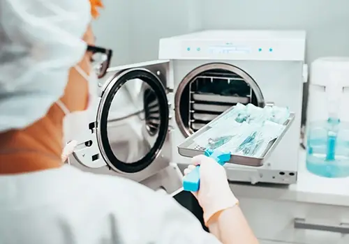 Healthcare worker places a tray of instruments into a tabletop autoclave for sterilization, PPE visible around.