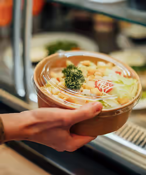 Person's hand holding a round container with vegetable soup and parsley garnish at a cafeteria counter.