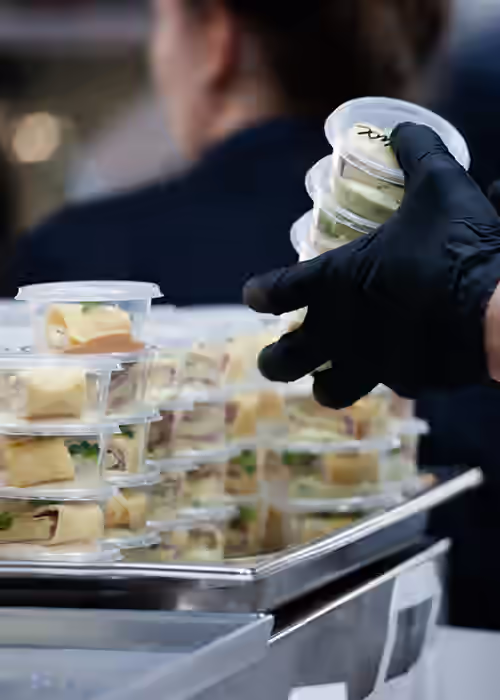 Caterer in gloves arranging small plastic cups of bite-sized foods on a stacked serving tray at a catered event.
