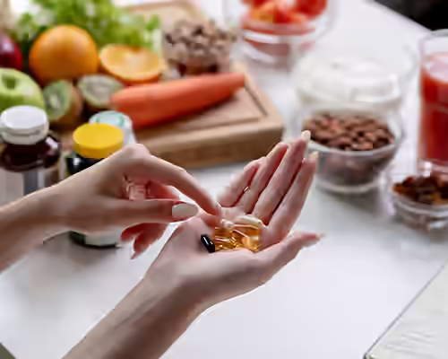 Close-up of hands holding gel capsules in a bright kitchen, with jars of pills and fresh fruit visible on the counter.