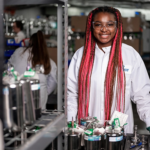 Smiling woman in a white lab coat with pink braids at a lab workstation beneath metal equipment.