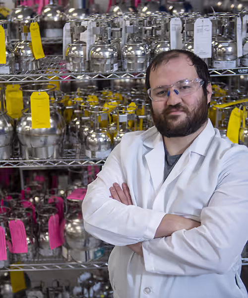 Male scientist in a white lab coat with arms crossed in a lab, shelves of tagged metal vessels behind him.