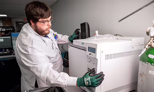 Lab technician in a white coat and green gloves adjusts a large analytical instrument in a lab setting, monitoring controls.