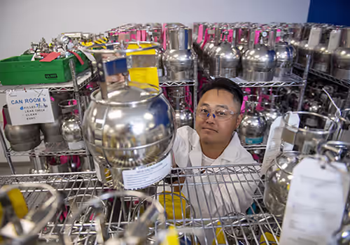 Lab technician wearing safety goggles handles a large metal gas cylinder among racks of cylinders in a lab storage area.