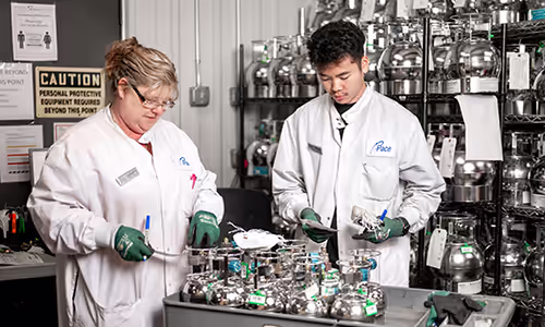 Two lab technicians in white coats and gloves work on stainless steel equipment at a lab bench in a busy lab environment.
