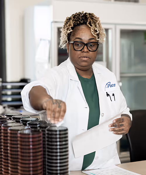 Black woman in a lab coat organizing stacked sample vials on a lab countertop. Pace® Scientist working in Legionella Laboratory.